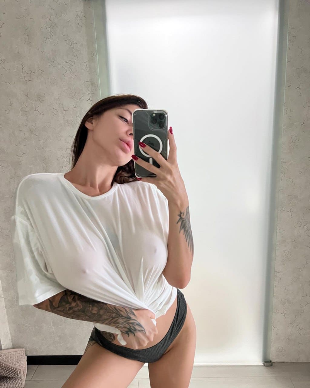 A woman takes a mirror selfie in a white t-shirt pulled up to reveal dark underwear, holding a phone that covers her face.
