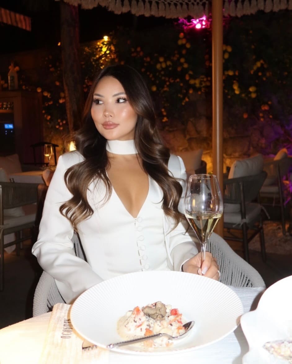 At a restaurant table, a woman in a white high-neck top holds white wine, with a plate of food in front.