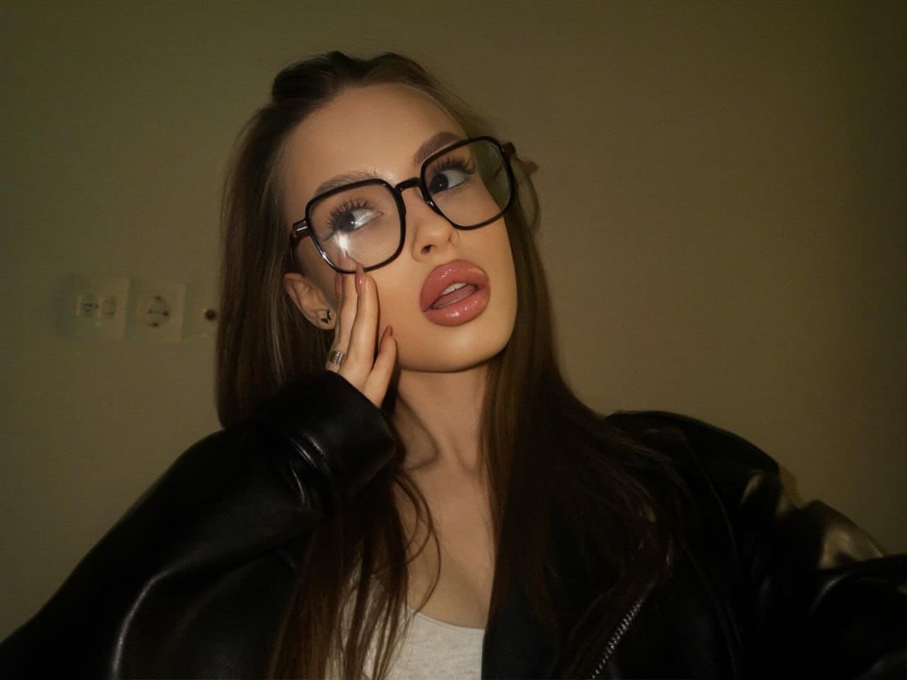 A woman with glasses and a black leather jacket poses indoors, resting a hand on her cheek against a plain wall with outlets.