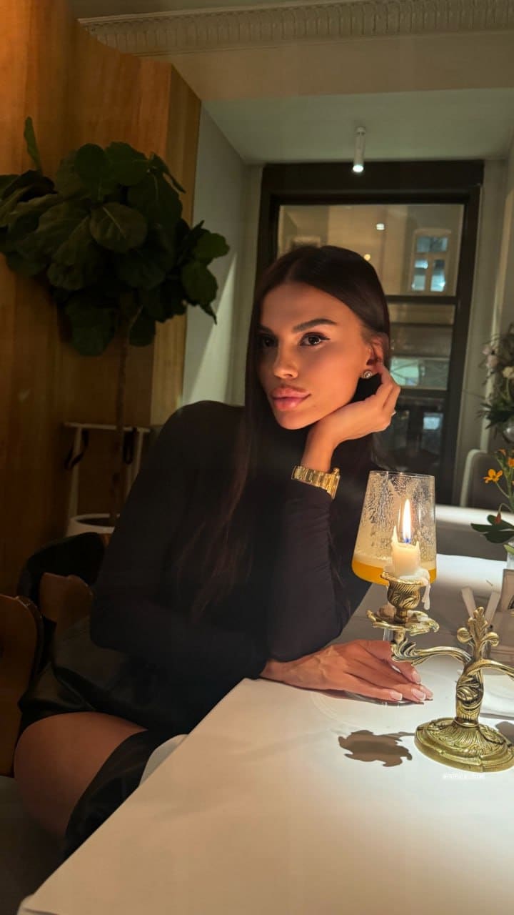A woman in a black long-sleeved top rests her chin on her hand at a table with a lit candle and a glass of orange juice.