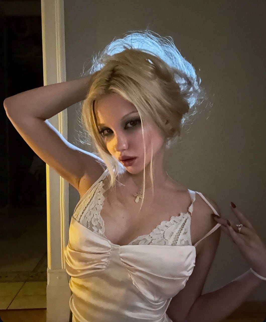 A blonde woman with hair up stands indoors in a cream satin camisole, one hand behind her head and the other on her shoulder.