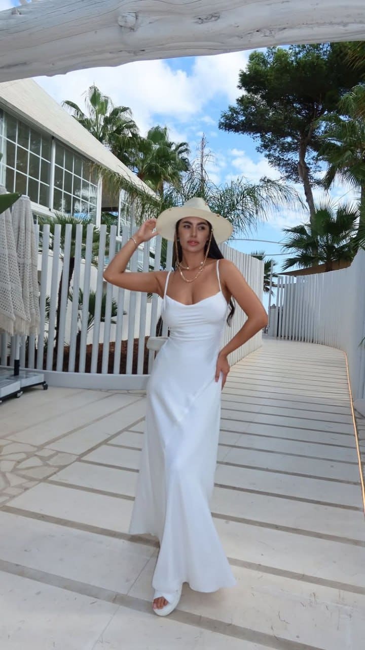 A woman in a long white dress and wide-brimmed straw hat stands on a tiled walkway, touching her hat brim, with palm trees and white fencing behind.