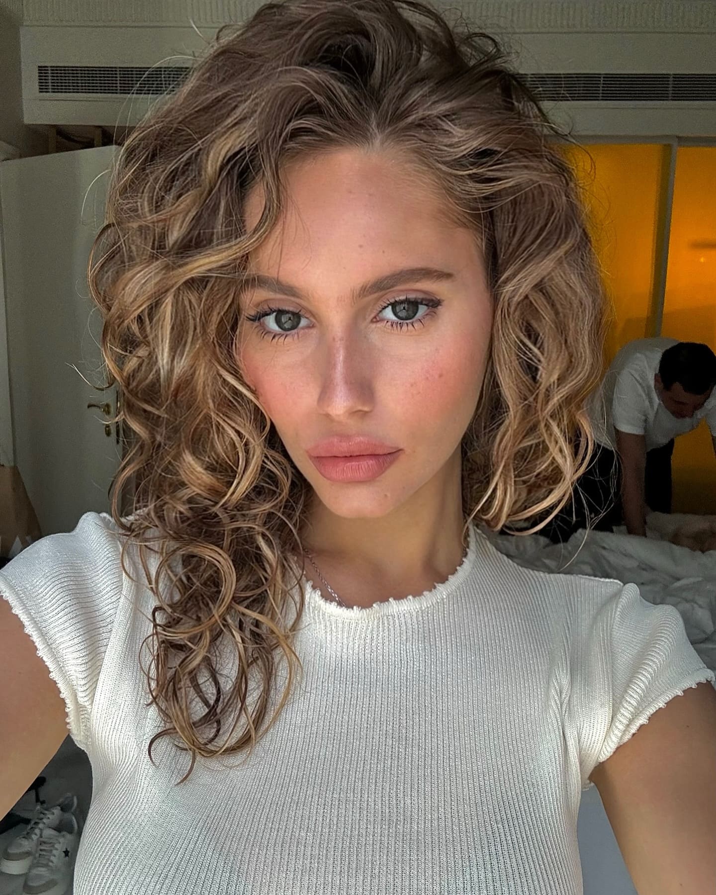 Mimi takes a selfie in a white ribbed top, with a bed and another person visible in the background.