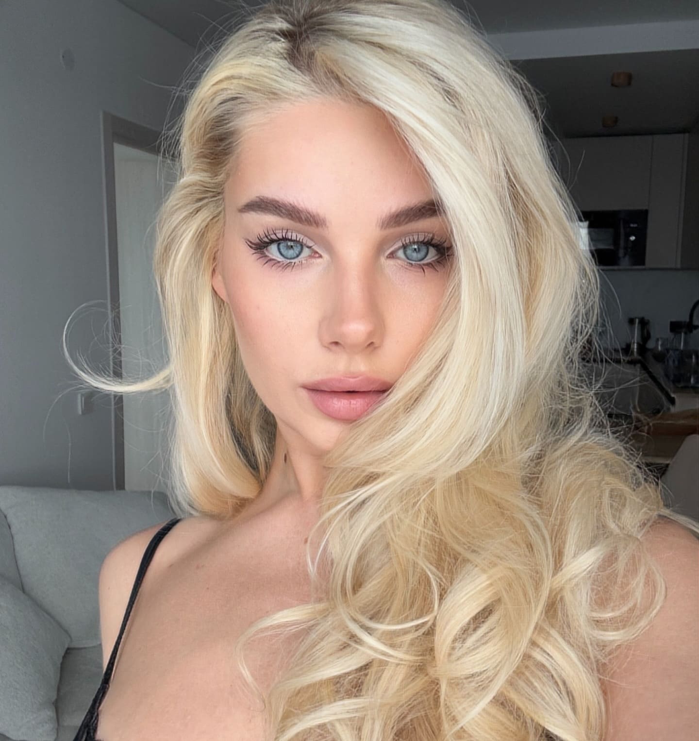 Selfie of a platinum-blonde woman in a black spaghetti-strap top, posed indoors near a grey sofa and kitchen.