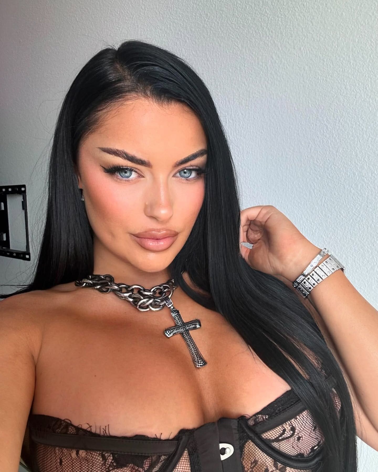 Zira poses in a black lace strapless top and chain necklace against a plain light wall.