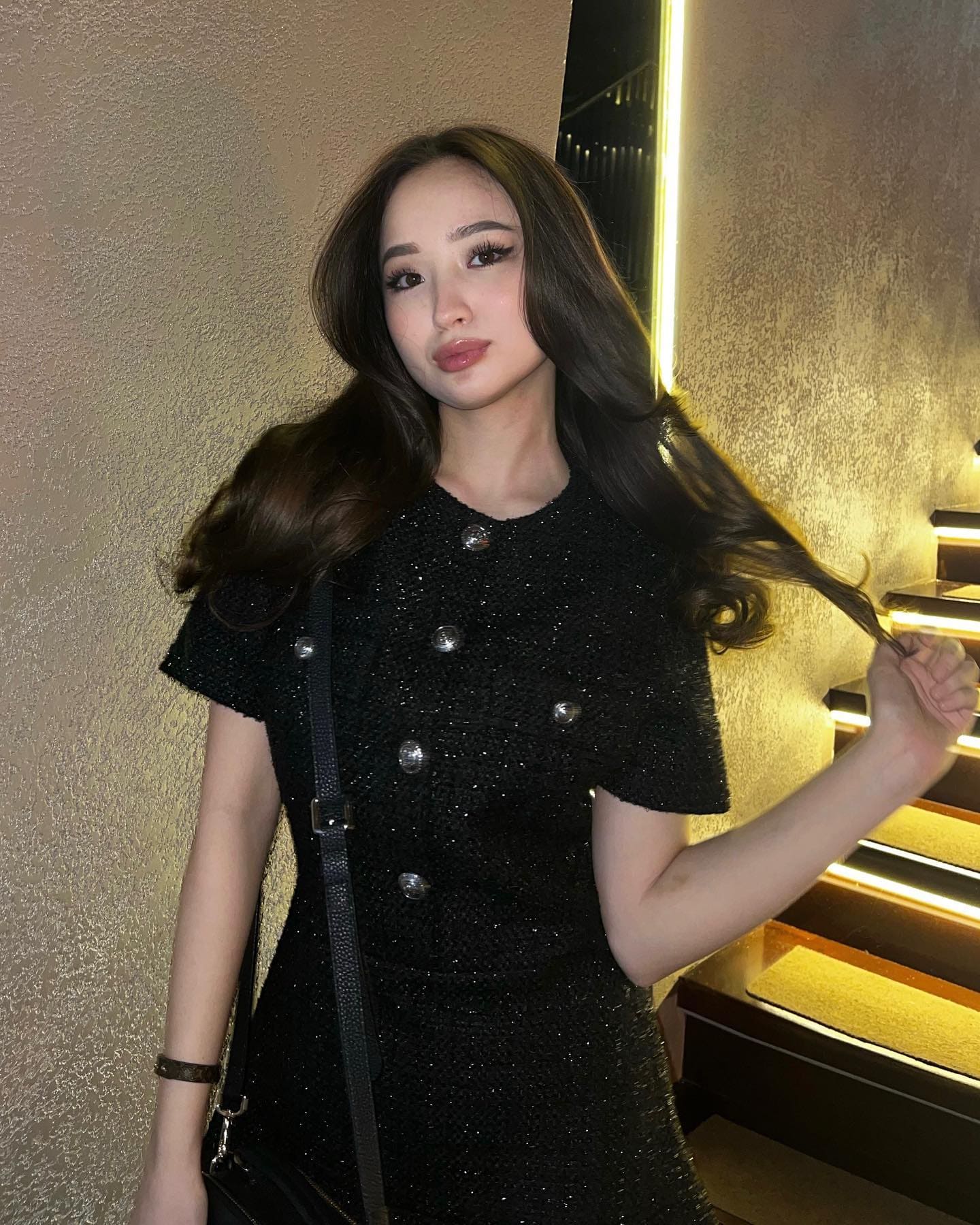 Clarensa stands indoors by a staircase in a black textured dress with silver buttons, holding a strand of her dark wavy hair.