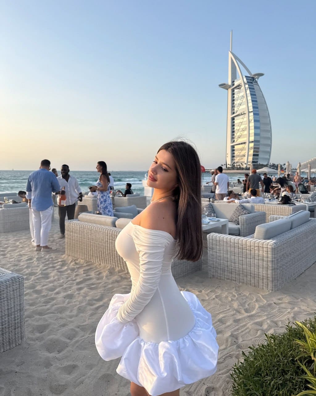A woman stands on a beach in a white off-the-shoulder mini dress, looking back over her shoulder near a sail-shaped building.