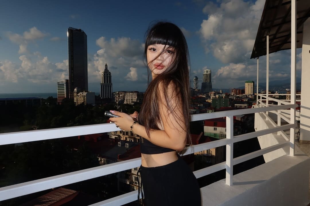 A woman in black crop top and pants looks back over her shoulder on a balcony, holding a phone, city skyline visible.