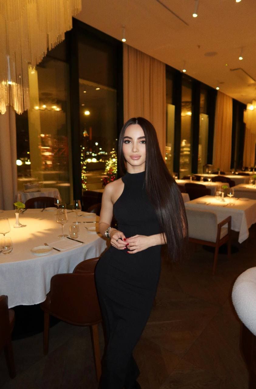 A woman with long dark hair stands in a restaurant wearing a sleeveless black high-neck dress, hands clasped near a set table.