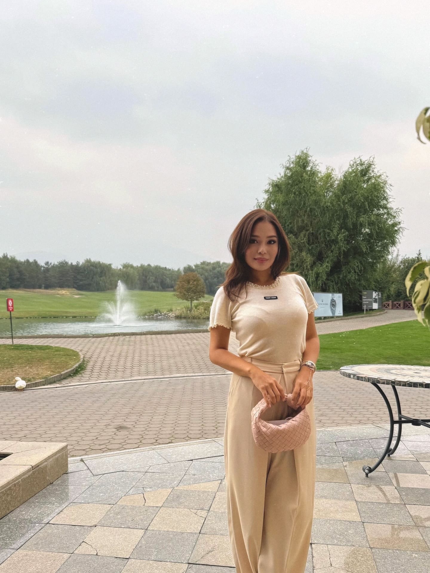 A woman stands on a paved patio in a beige top and trousers, holding a woven bag, with a fountain and green lawn visible under an overcast sky.