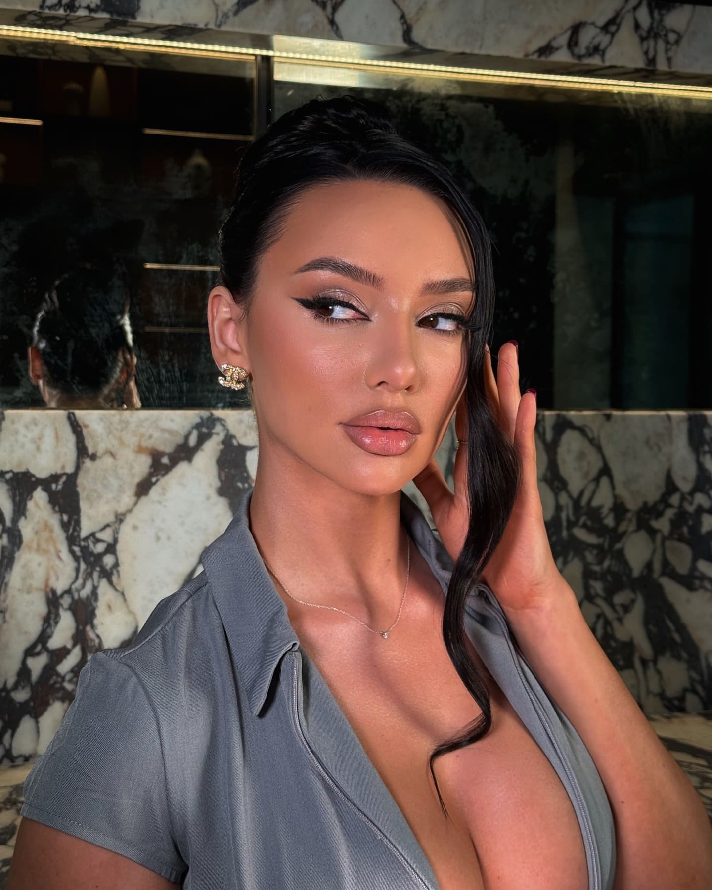 Jina with dark hair pulled back wears a grey short-sleeved top with an open neckline, posing near a marble wall and mirror.