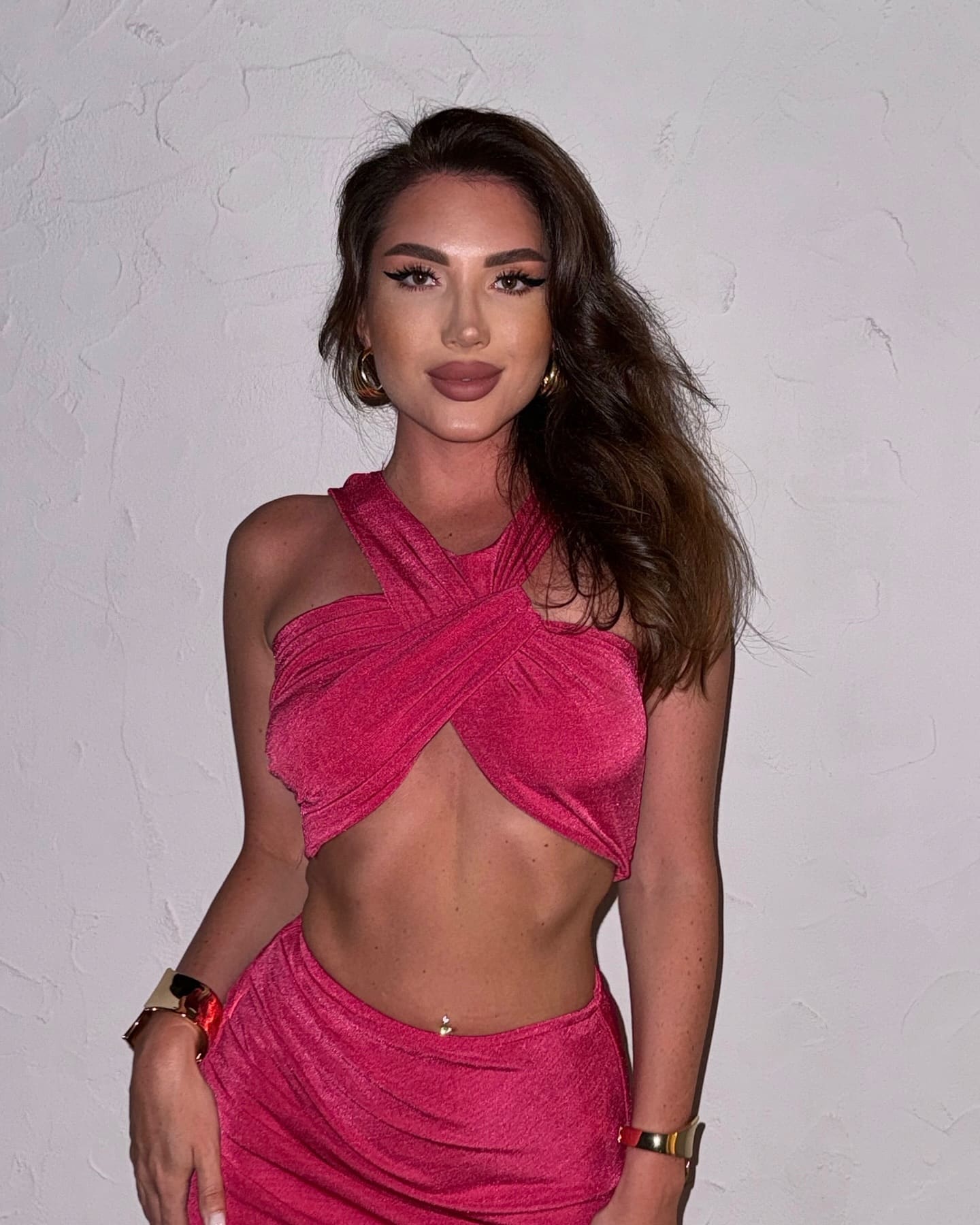 Malika stands against a white wall in a pink crisscross halter top and high-waisted skirt, wearing gold hoop earrings and wide bracelets.