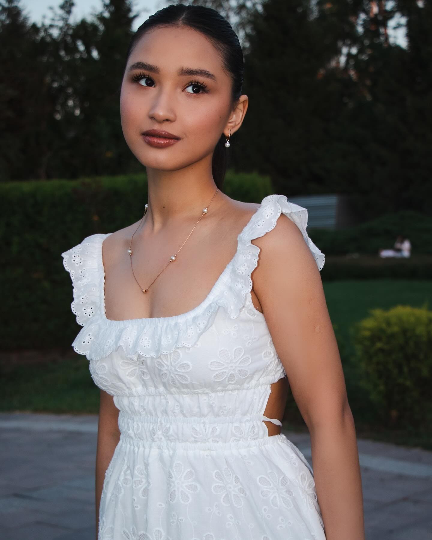 Mariel stands outdoors before green hedges, wearing a white eyelet dress with ruffled straps and a square neckline.