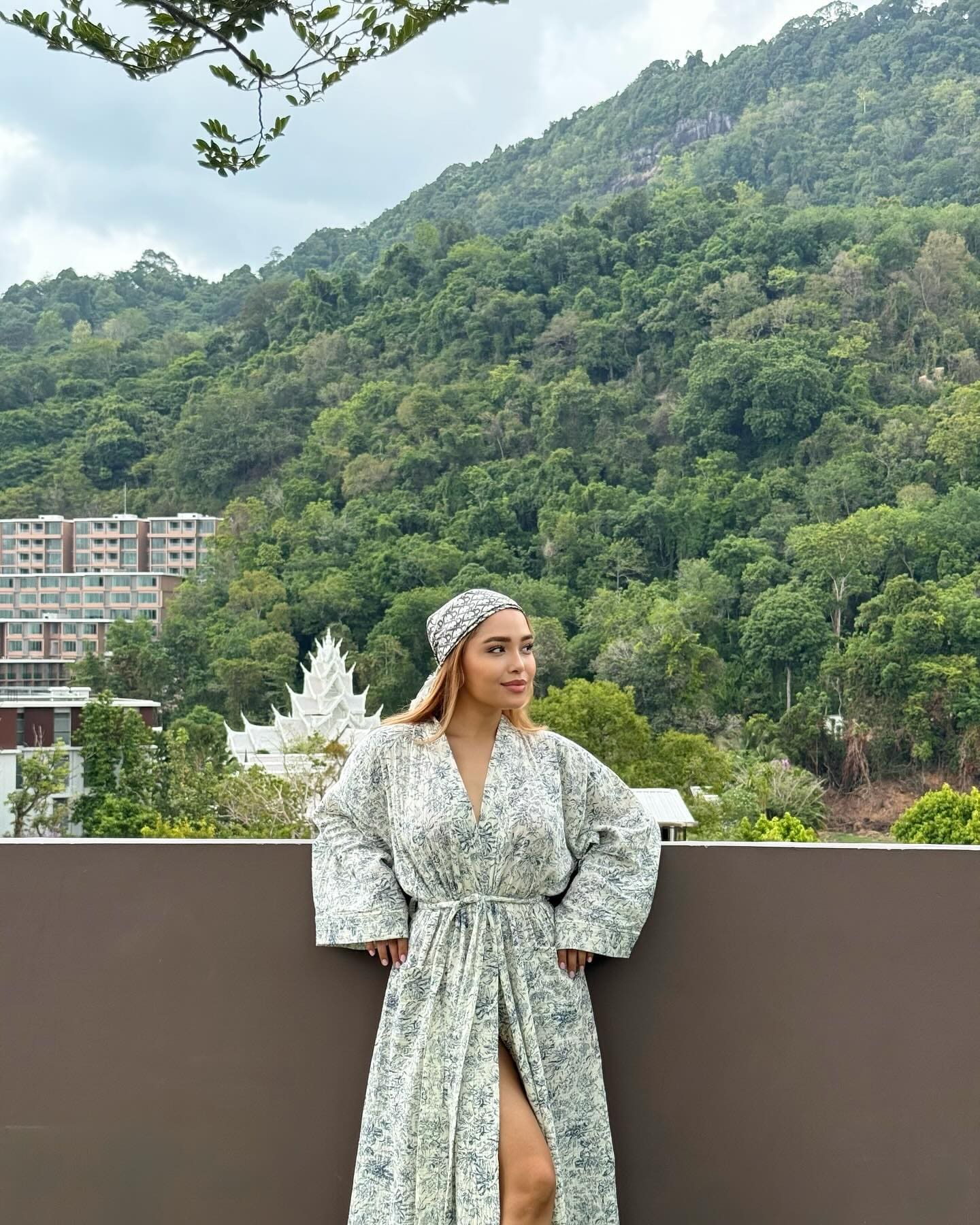 A woman stands on a balcony in a floral robe and headscarf, hands on hips, with a green hillside and white temple structure in the background.