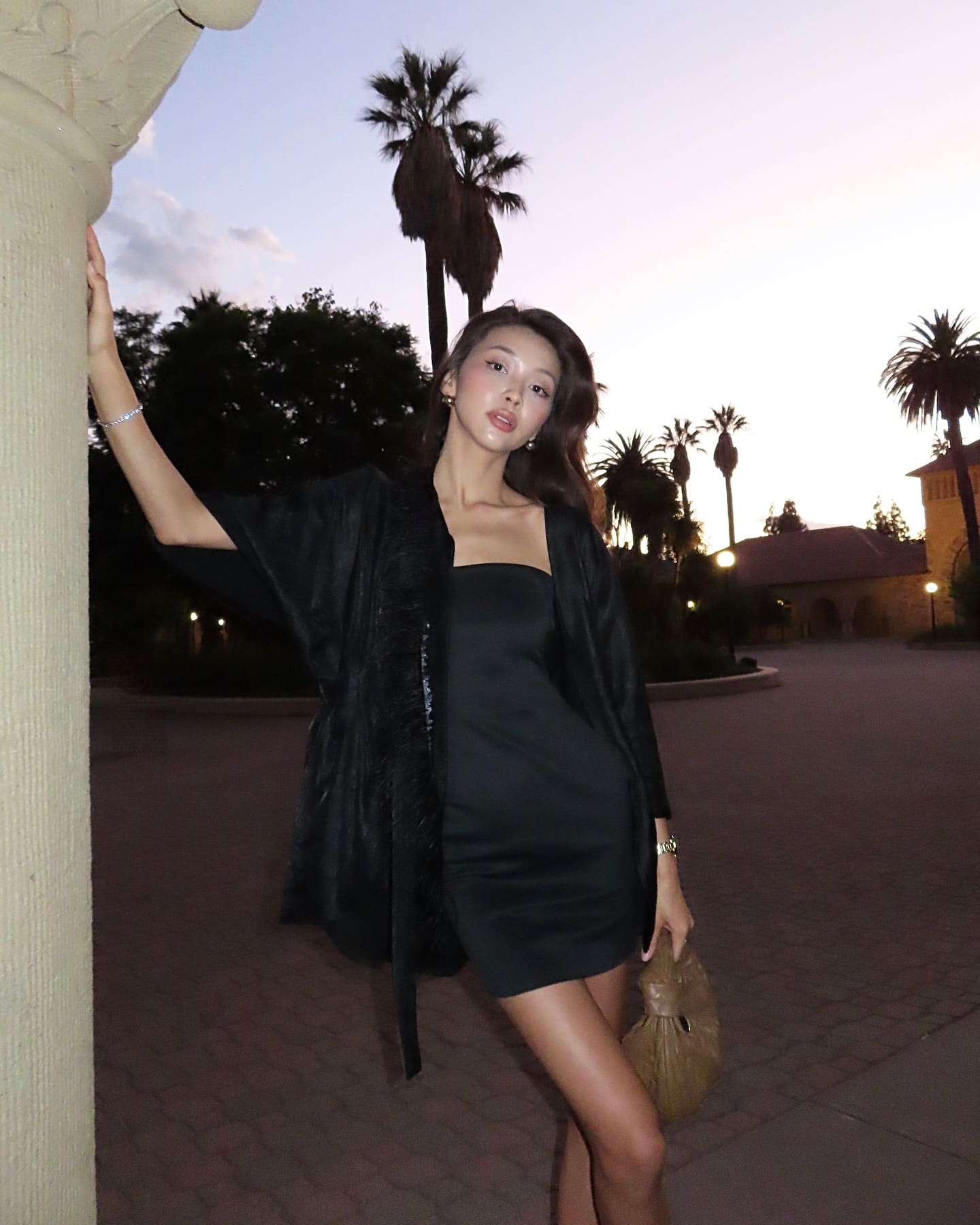At dusk outdoors, she wears a black strapless mini dress and open jacket, resting a hand on a stone column with palm trees behind her.