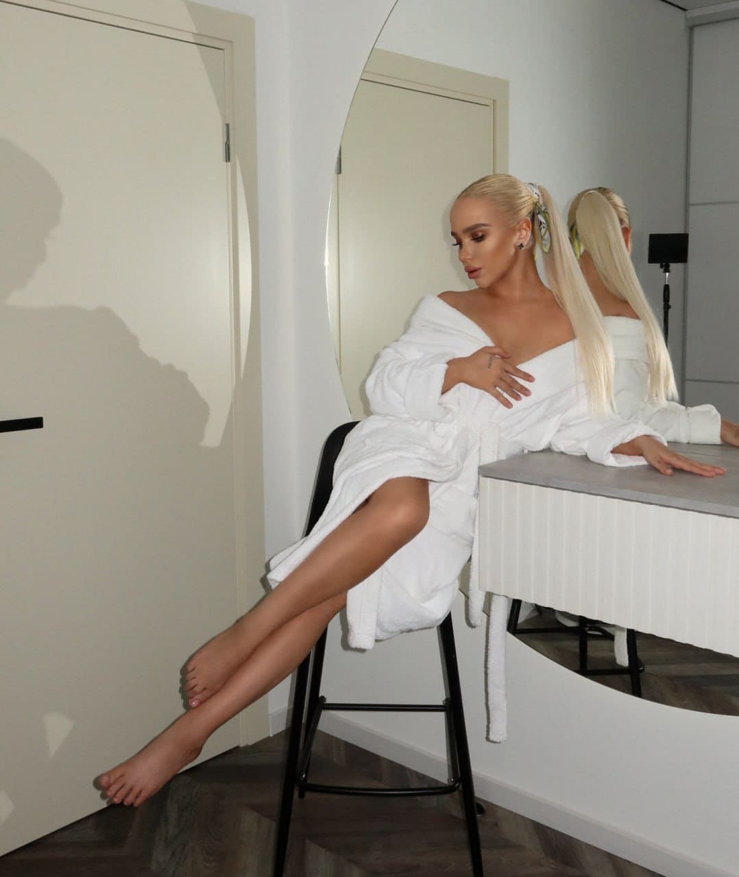 A blonde woman with a high ponytail sits barefoot on a black stool in an open white bathrobe, in a room with a large mirror and counter.