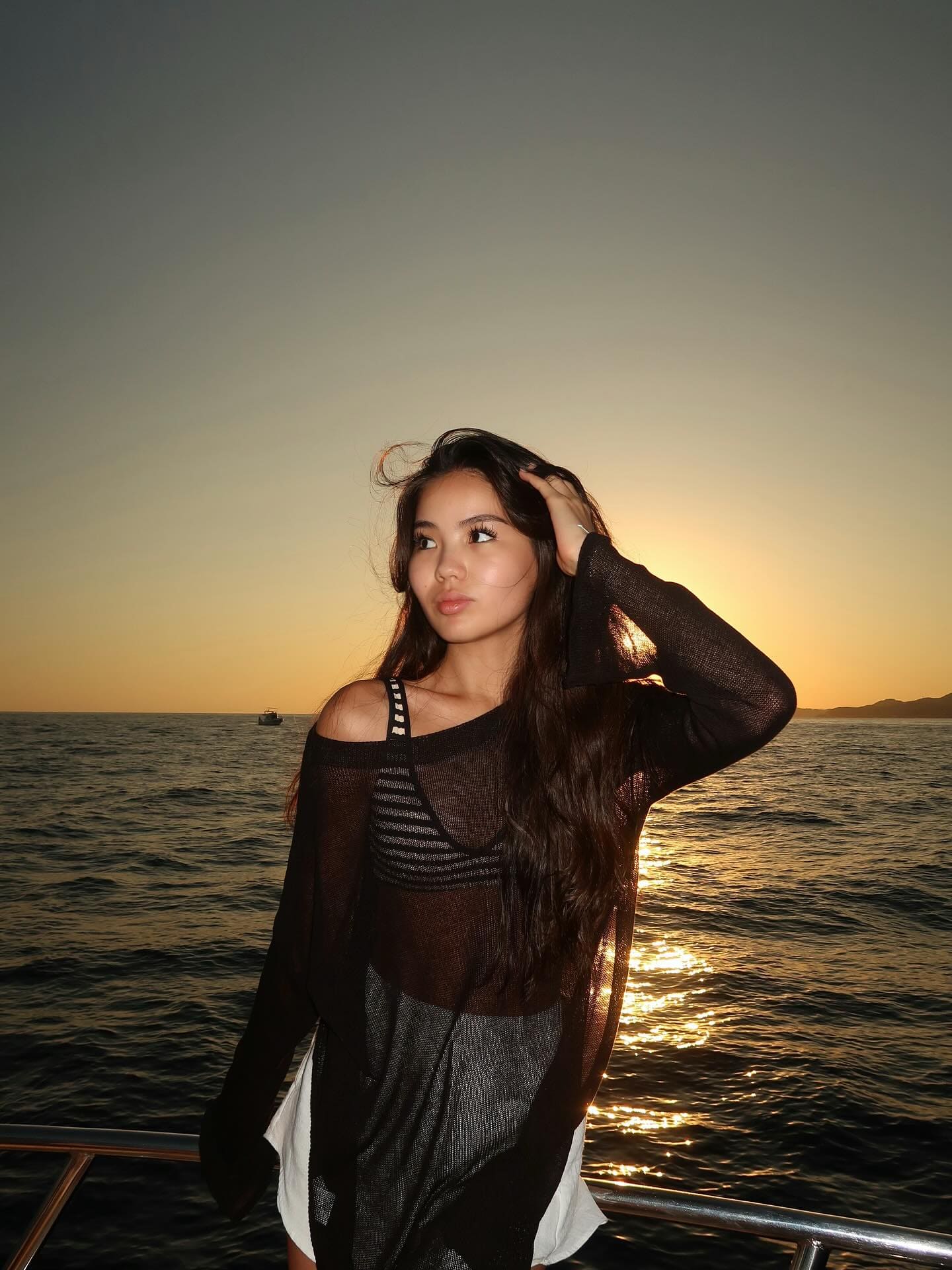 A woman on a boat at sunset wears a sheer black top over a striped tank and white shorts, with one hand raised to her hair.