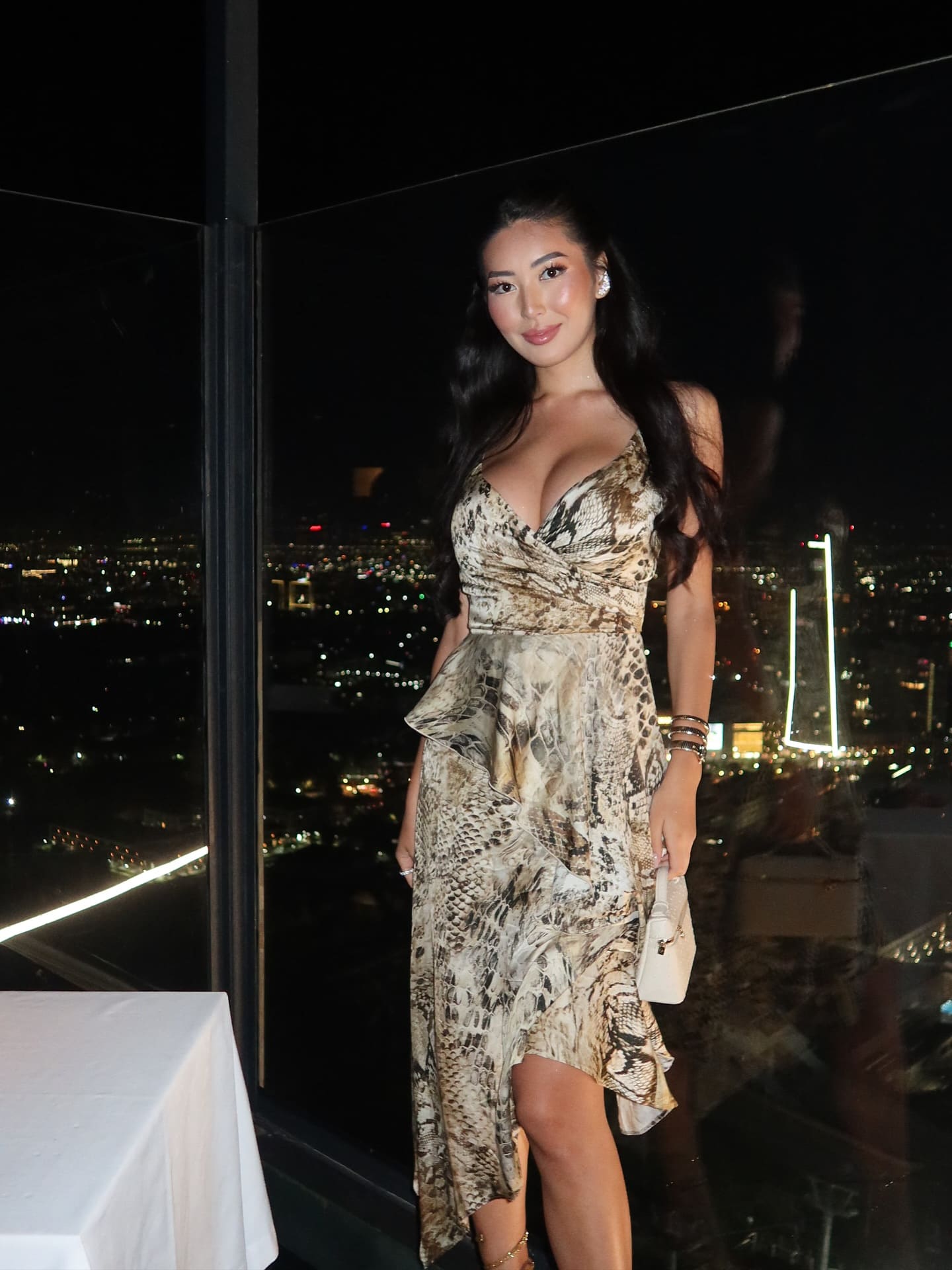 A woman in a snake-print dress holds a white handbag while standing by a window overlooking a city skyline at night.