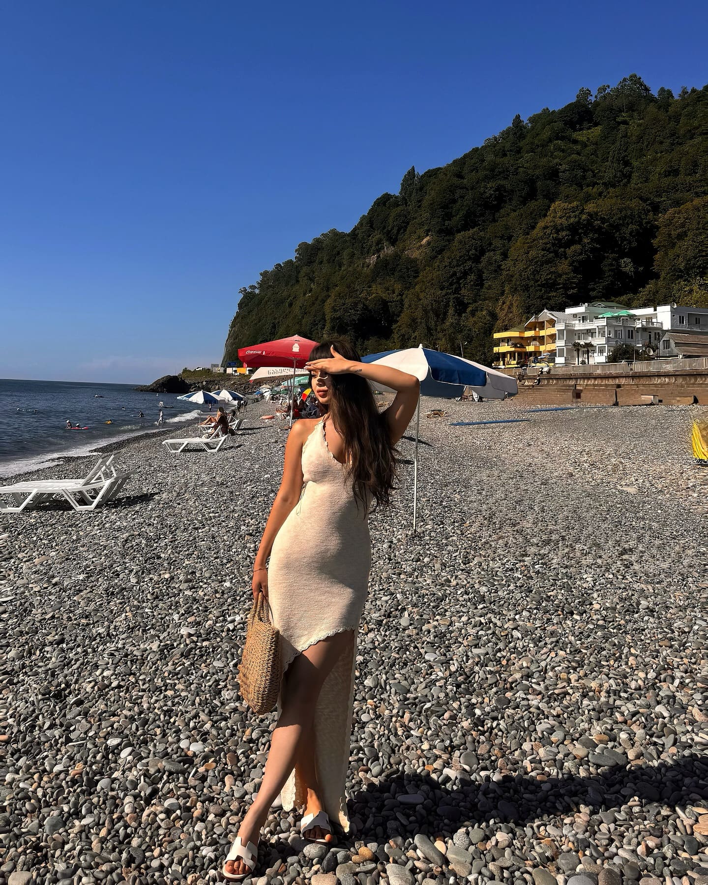 A woman in a cream slit dress holds a woven bag and shades her eyes on a pebble beach with umbrellas and hills behind.