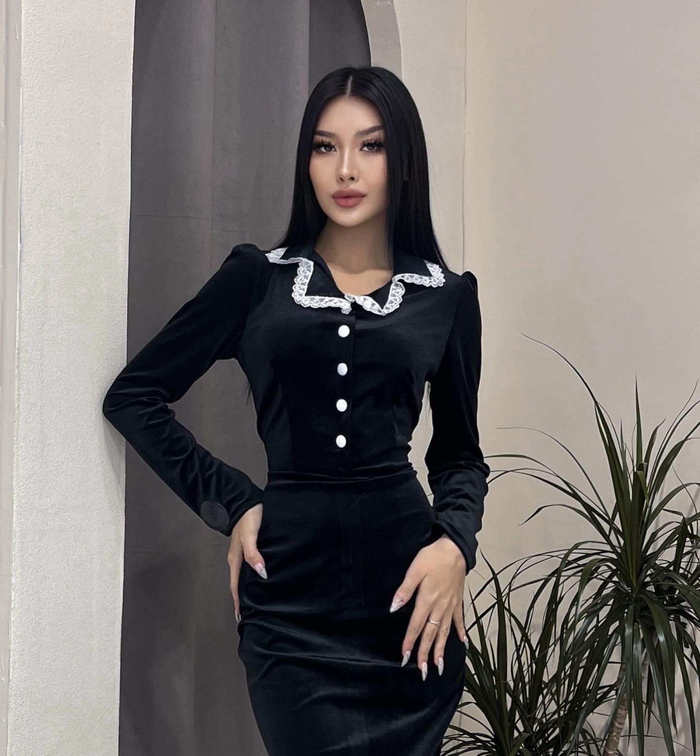 Evangeline stands with hands on hips in a black velvet dress with a white lace collar, before a textured wall and potted plant.