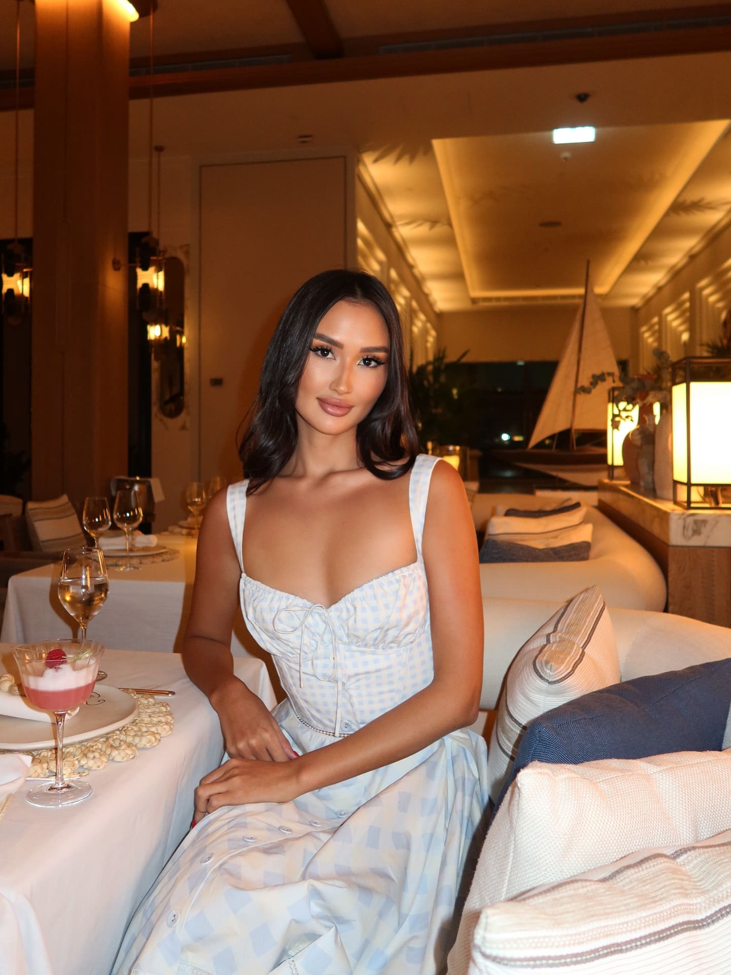 Jasmina sits at a restaurant table in a blue and white gingham dress, hands on lap, with dessert and wine in front.