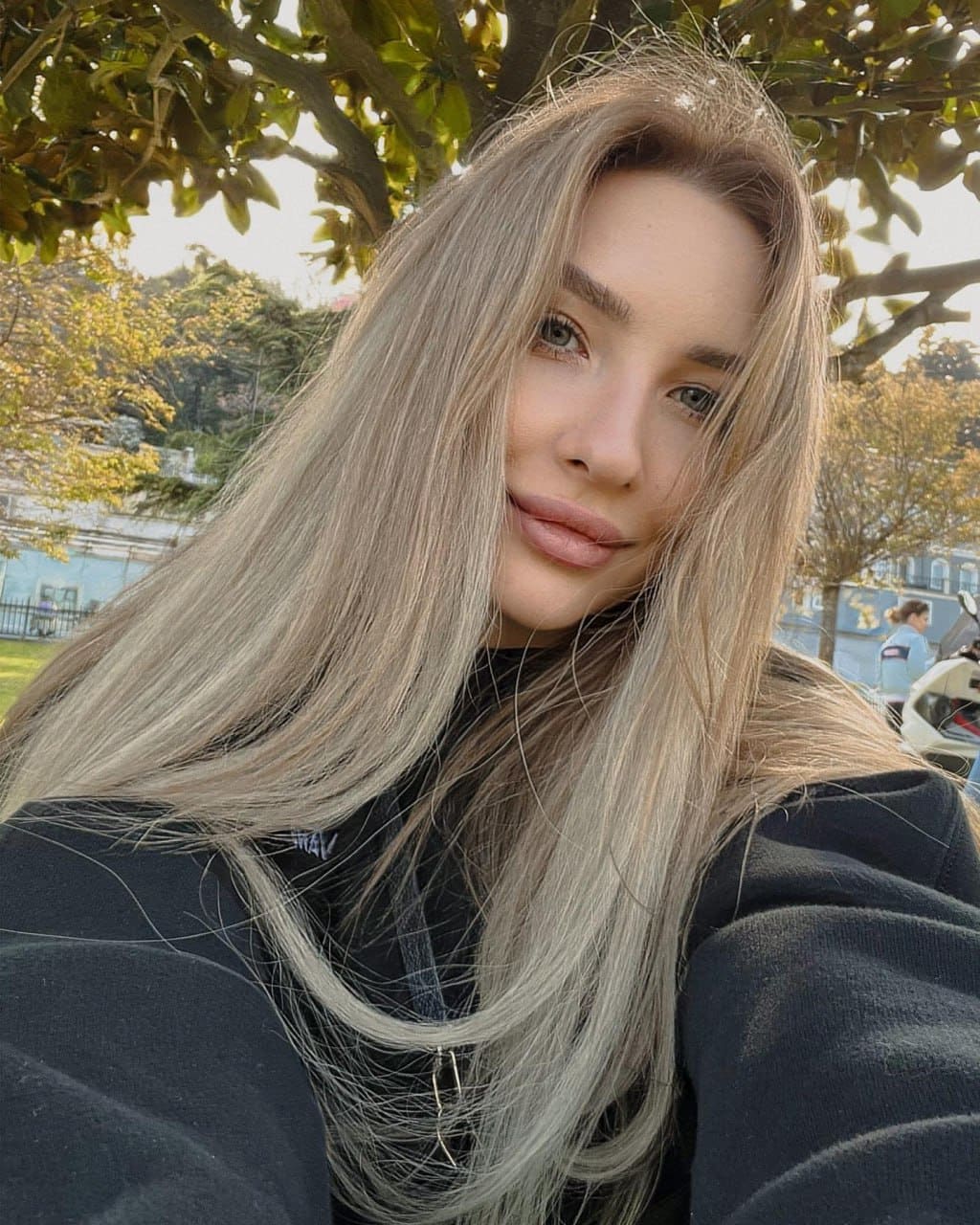 Sofia takes a selfie outdoors in a dark hooded sweatshirt, with trees and a park-like setting behind her.