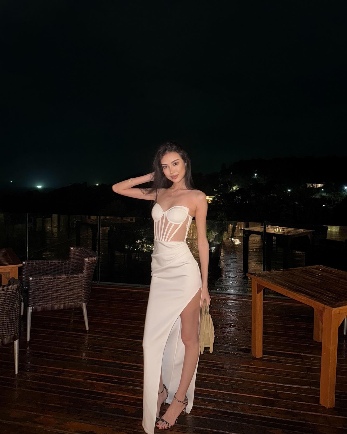 A woman stands on a wet deck at night in a white cutout dress, one hand in her hair, near wicker chairs and a dark background.
