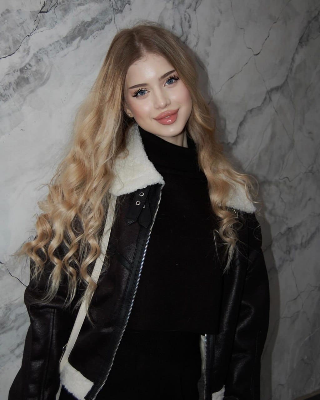 Lily stands against a grey marble wall wearing a black turtleneck under a black leather jacket with a white shearling collar.