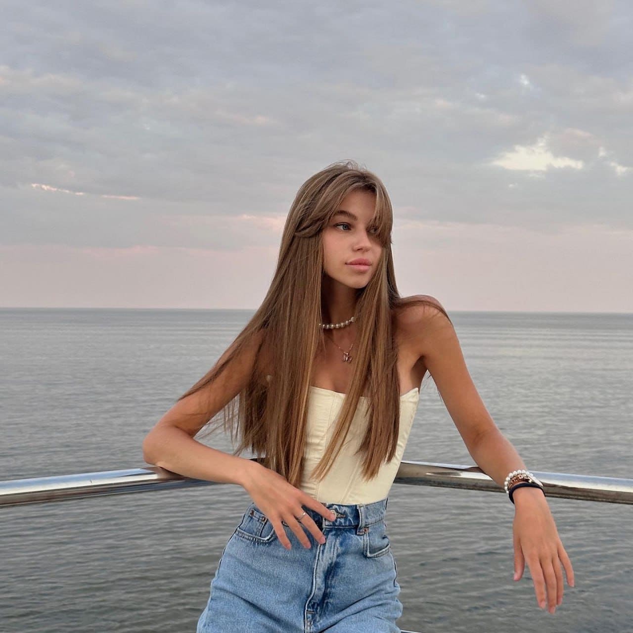 Renata stands by a railing near water in a white strapless top and jeans, with a cloudy sky behind her.