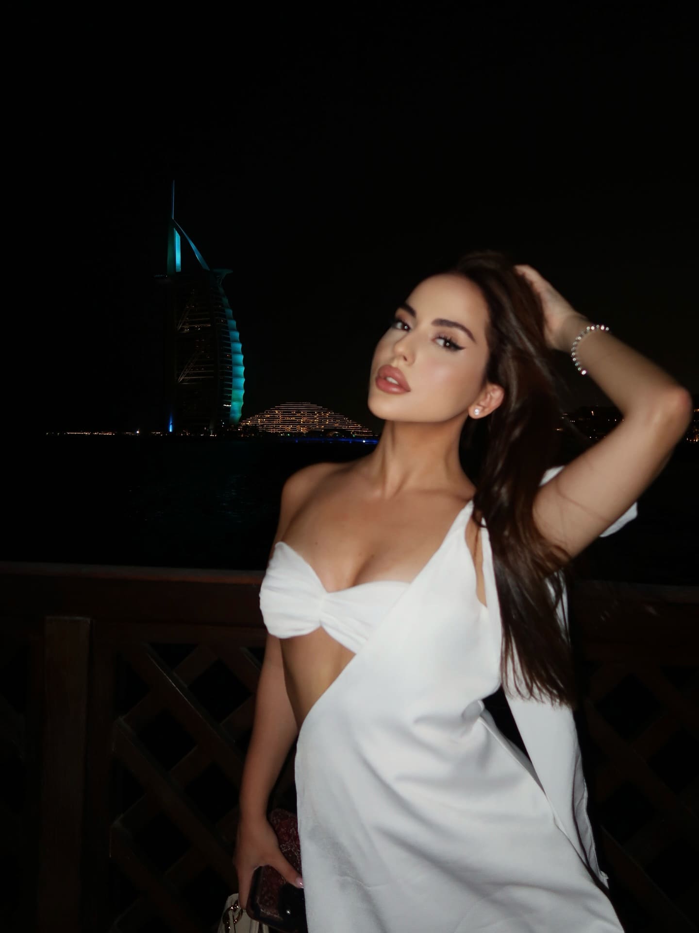 At night, a woman in a white one-shoulder dress with a waist cutout stands by a wooden railing, with a lit sail-shaped building behind.