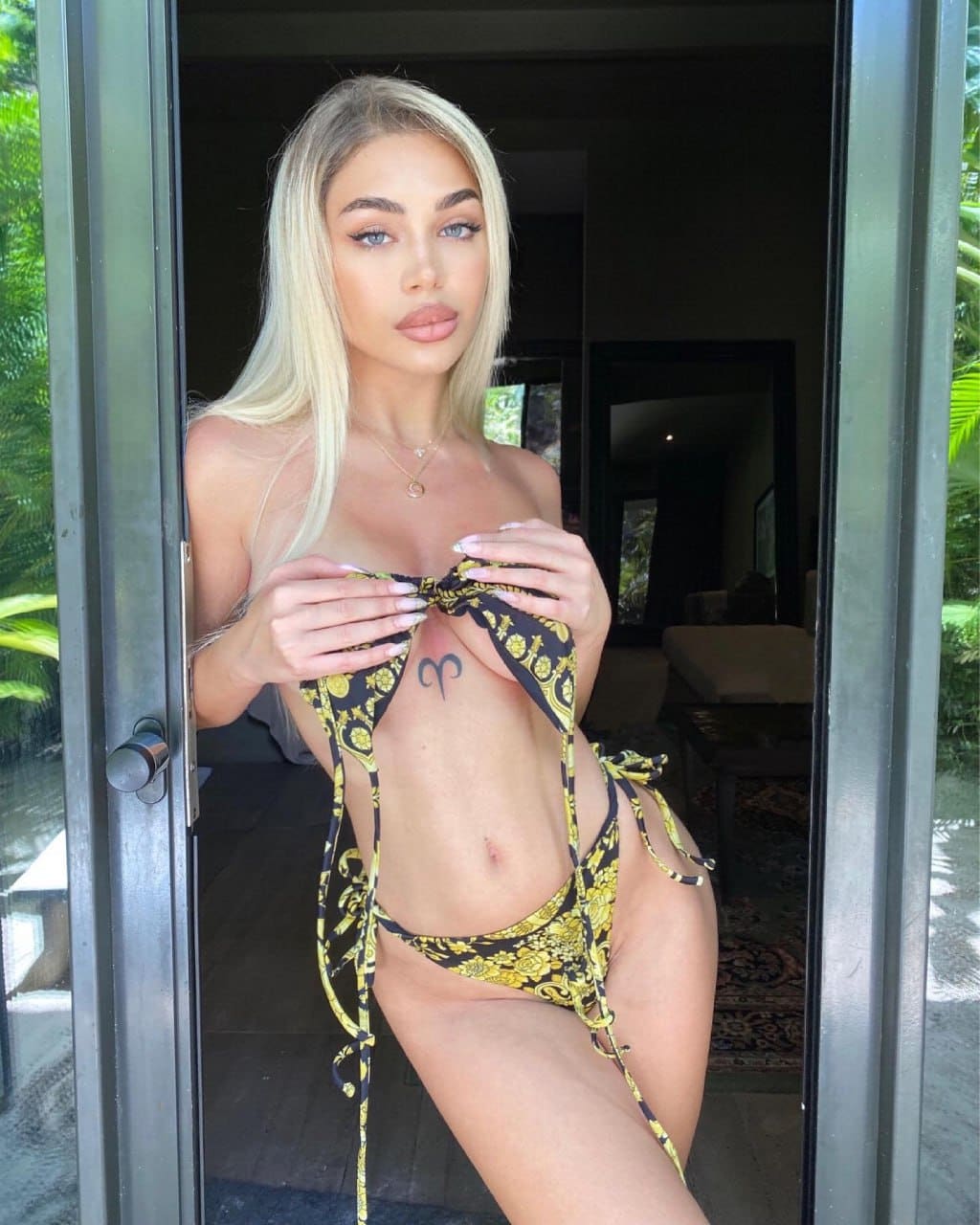 A blonde woman stands in a doorway wearing a black and gold patterned string bikini, holding the straps together with both hands.