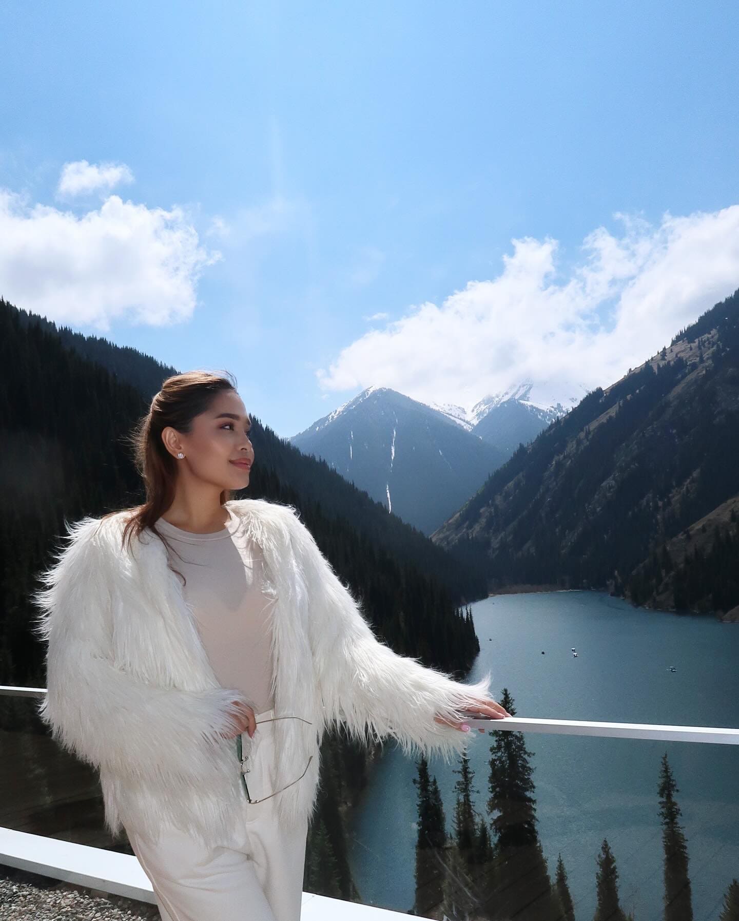 A woman in a white fluffy coat and pants stands at a railing overlooking a mountain lake, holding glasses in one hand.